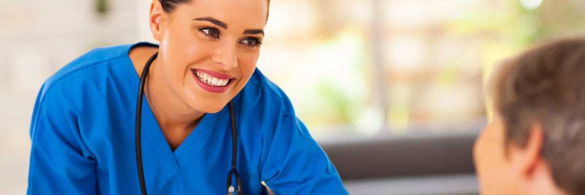 Zdjęcie przedstawia uśmiechniętą pielęgniarkę pochylającą się nad siedzącą pacjentką.