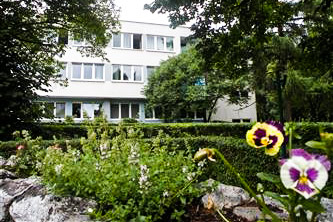 Zdjęcie budynku Miejskiego Centrum Opieki w Krakowie. Budynek jest otoczony przez zieleń, na pierwszym planie widać kwiaty.